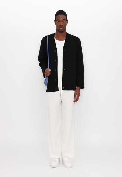 Black button-up jacket with a relaxed fit, worn over a white top, paired with cream pants and white sneakers, holding a blue shoulder bag.