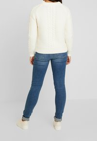 Person som står vänd bort wearing a white cable-knit sweater, blue skinny jeans med manschetter och vita sneakers mot en enkel bakgrund.