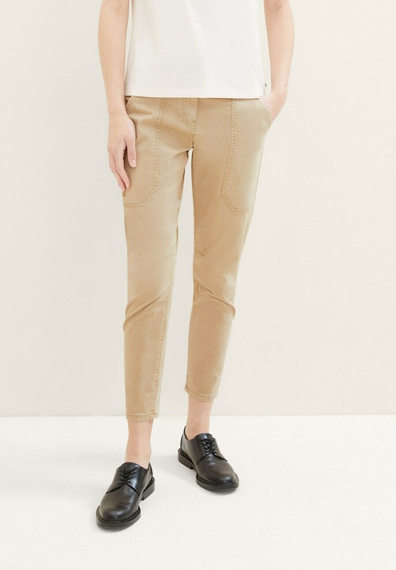 Beige tapered pants with pocket details and stitching accents, paired with black leather lace-up shoes. Smooth texture and fitted design.