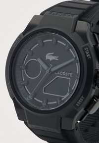 Zwarte Lacoste-horloge met ronde wijzerplaat, zilveren uurmarkeringen, drie wijzers en zwarte rubberen strap met getextureerde zijkanten.