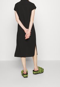 Czarna sukienka z krótkimi rękawami i bocznym rozcięciem, połączona z klapkami w kolorze neonowej zieleni w czarno-białe paski zebra na jasnoszarym tle.