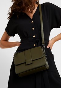 Mujer con un vestido negro abotonado que lleva un bolso cruzado de cuero verde oliva con tapa y correa ajustable.