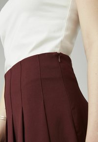 Jupe plissée bordeaux en tissu lisse, dotée d'une fermeture éclair sur le côté, associée à un haut blanc ajusté. Gros plan sur la taille.