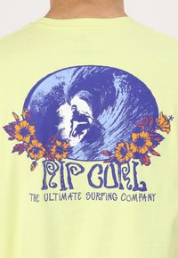 Šviesiai geltonas marškinėliai su mėlyna ir oranžine grafika, vaizduojančia banglentininką bangos viduje, apsuptą gėlių akcentų, ir užrašu "Rip Curl".