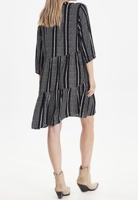 Mustat ja valkoiset raidalliset mekko, jossa on kolmivartiset hihat, kerrostettu helma, valmistettu kevyestä kankaasta. Näkyvissä on myös beiget nilkkasaappaat.