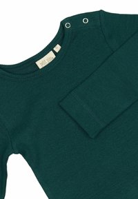 Indumento a maniche lunghe di un verde scuro con una texture a coste, dotato di bottoni a pressione sulla spalla e una scollatura rettangolare. Etichetta visibile all'interno.