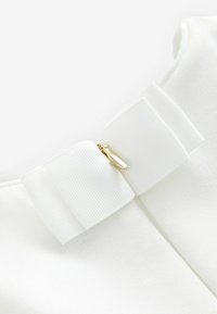 Weißer Stoff mit strukturiertem Finish, der mit einer markanten, breiten weißen Schleifenbinde und einem goldenen Reißverschlussdetail auf der Rückseite versehen ist.