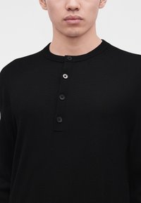Homme portant une chemise Henley noire à manches longues avec quatre boutons, un argenté et trois noirs, sur un fond uni gris clair.