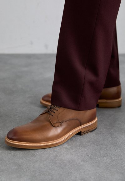 Hnedé kožené šnurovacie topánky s okrúhlou špičkou, kontrastným svetlým prešívaním a béžovou podrážkou, v páre s tmavo bordovými nohavicami.