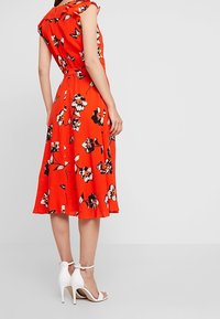 Robe fleurie rouge avec manches courtes à volants, taille ceinturée et jupe longueur genou. Motifs floraux noirs et blancs. Escarpins blancs à bride de cheville.