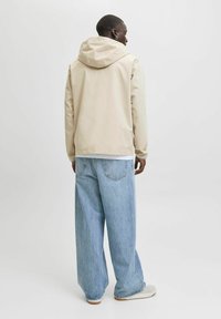 Chaqueta impermeable beige con capucha, corte relajado y mangas largas. Combinada con jeans de pierna ancha azul claro y zapatos grises.