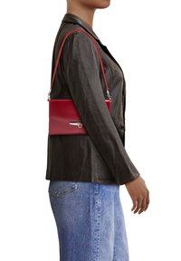 Borsa a tracolla rossa in pelle con un design sottile, tracolla regolabile e dettaglio della chiusura in argento. Indossata al fianco di un modello sopra una giacca nera.