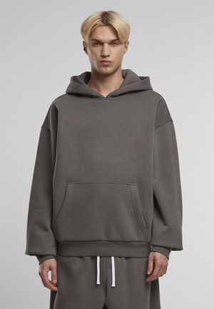 Szara bluza z kapturem, wykonana z miękkiej tkaniny, z kieszenią z przodu, ściągaczami na rękawach i przestronnym kapturem. Białe sznurki podkreślają jej design.