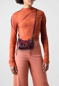 Liten burgunderrød crossbody-veske med en teksturert overflate, kjedehank og dekorative nagler, kombinert med en oransje langermet topp og rosa bukser.