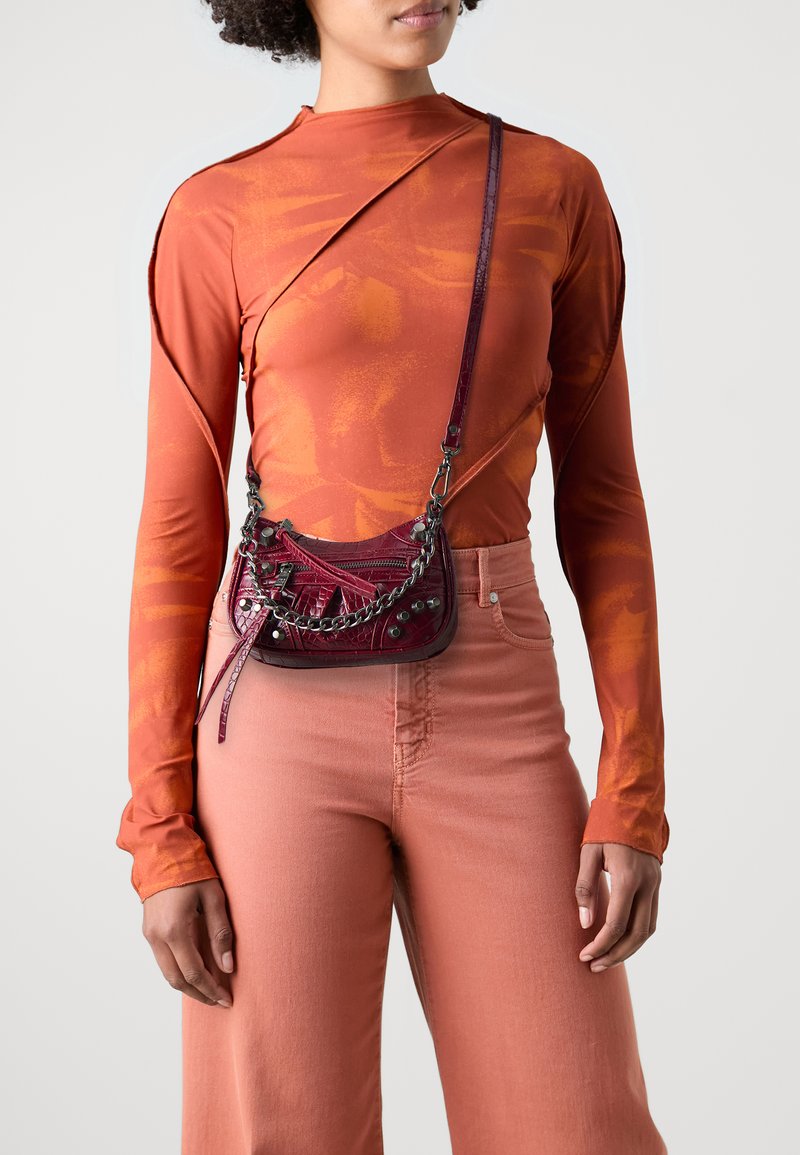 Liten burgunderrød crossbody-veske med en teksturert overflate, kjedehank og dekorative nagler, kombinert med en oransje langermet topp og rosa bukser.