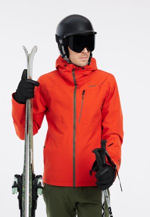 Orange waterproof ski jacket with hood, zippered pocket, paired with black gloves and ski poles. Matte black helmet and large dark goggles.
