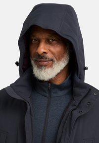 Veste à capuche navy avec fermeture éclair et accents à pression, dotée d'une doublure intérieure douce et d'un col haut, portée sur un haut en polaire bleu foncé.