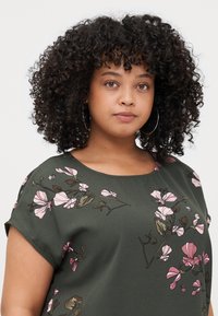 Green short-sleeve top featuring light pink floral patterns. Smooth fabric with a crew neckline and relaxed fit design.