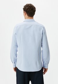 Camisa de botones azul claro con mangas largas, tela texturada, cuello puntiagudo y dobladillo redondeado, con puños abotonados y pinzas en la espalda.