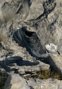 Schwarzer Sportschuh mit strukturiertem Obermaterial, robuster Sohle und verstärktem Zehenbereich, schwebend über felsigem Terrain und das elegante Design präsentierend.