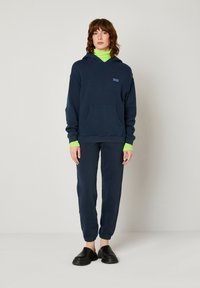Tumši zili hoodie ar priekšējo kabatu, kam ir neliels logo. Kombinēts ar atbilstošiem biksēm un melniem slip-on apaviem. Zaļš apkakles akcents.