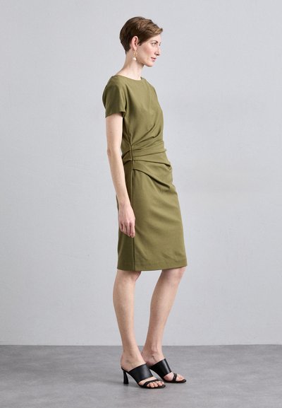 Robe ajustée vert olive avec des manches courtes, détails drapés sur le côté et une longueur jusqu'aux genoux. Associée à des talons ouverts noirs.