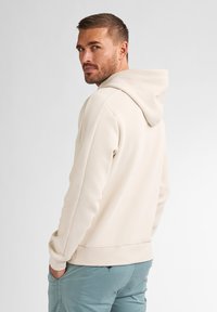 Beige Kapuzenpullover mit verstellbarem Kapuzenzug, enganliegendem Bündchen und angenehmem Material, kombiniert mit hellblauen Hosen, von hinten gezeigt.
