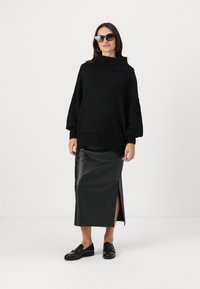 Pull en maille noir épais avec un col montant, associé à une jupe midi en cuir noir avec une fente sur le côté. Le modèle porte des lunettes de soleil surdimensionnées et des mocassins.