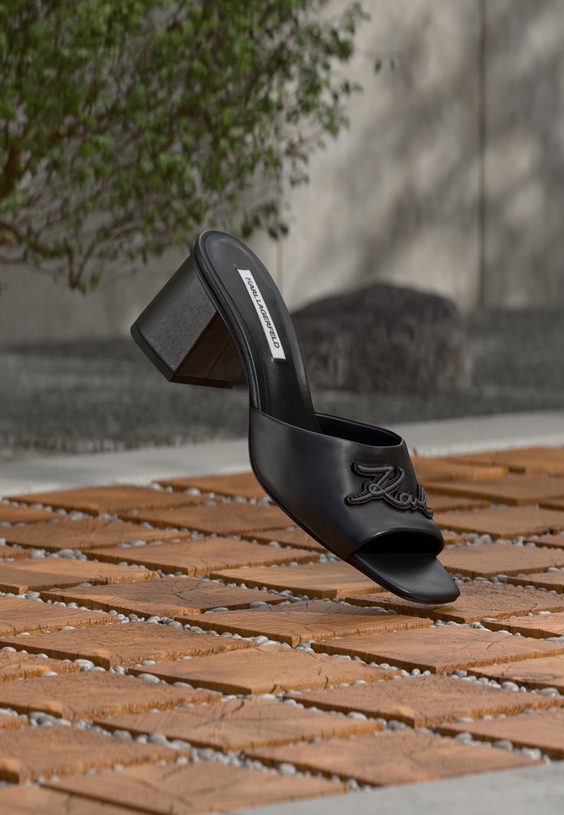 Μαύρο mule ανοιχτής μύτης με χοντρό τακούνι και την επιγραφή "Karl" κεντημένη στο λουρί, τοποθετημένο πάνω σε πλακόστρωτο με ξύλινα πλακίδια, με θαμπό φόντο.