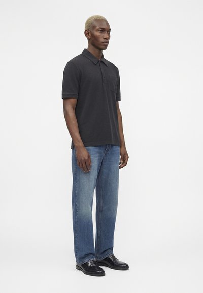 Άνδρας με κοντά ξανθά μαλλιά, φοράει μαύρο πόλο με κοντά μανίκια, μπλε τζιν παντελόνι με ίσια γραμμή και μαύρα δερμάτινα παπούτσια, στέκεται πλάγια.