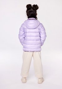 Giacca puffer lavanda con cappuccio, caratterizzata da texture trapuntata e polsini elasticizzati, indossata sopra pantaloni della tuta crema e sneaker bianche.