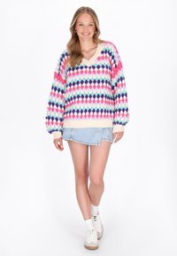 Tânără zâmbitoare, purtând un pulover colorat din croșetat, fustă mini din denim deschis, pantofi sport albi și cercei tip inel, stand pe un fundal alb.