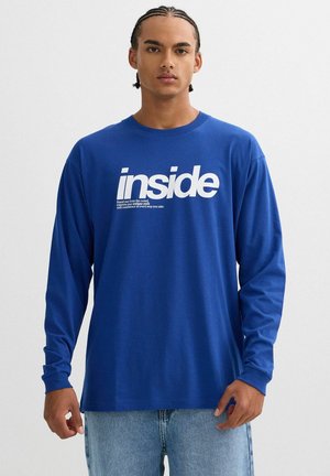 Camiseta de manga larga azul con texto blanco "inside" y texto adicional abajo. Fabricada con tela suave, con un escote redondo y un corte holgado.