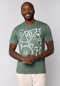 Groene katoenen t-shirt met een wit geometrisch ontwerp van fiets tandwielen, gereedschap en patronen. Model draagt een lichtbeige broek.