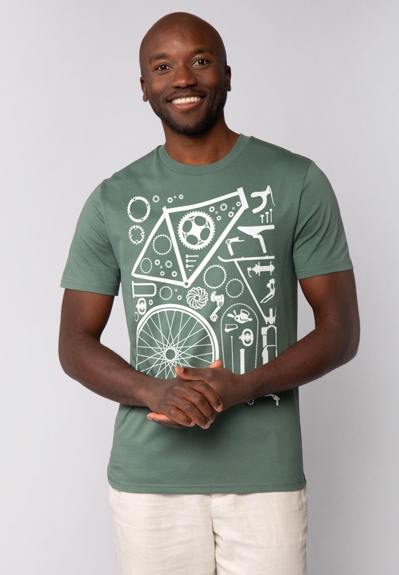 Groene katoenen t-shirt met een wit geometrisch ontwerp van fiets tandwielen, gereedschap en patronen. Model draagt een lichtbeige broek.