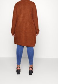 Cardigan lungo color ruggine in una lavorazione a maglia spessa, con scollatura rotonda e spacchi laterali, abbinato a jeans skinny blu e tacchi neri.