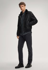 Giacca in shearling nera con colletto alto, chiusura a zip, polsini a costine, abbinata a jeans scuri e stivaletti neri.