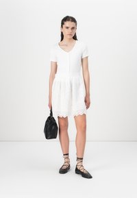 White dress with short sleeves, ribbed bodice, and a laser-cut lace skirt. Paired with black flat shoes and a textured black bag.
