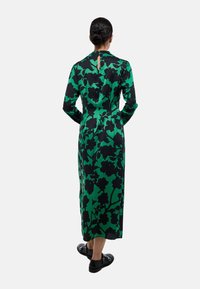 Robe multicolore avec un tissu vert à motif floral noir, des manches longues et un détail en trou de serrure dans le dos. Chaussures plates noires.