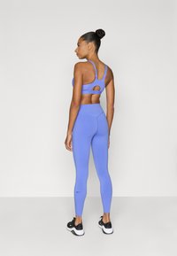 Μωβ αθλητικό σουτιέν με λεπτομέρεια κοψίματος και συμπληρωματικό κολάν με ψηλή μέση, φτιαγμένα από λείο, ελαστικό ύφασμα. Μαύρα αθλητικά παπούτσια ολοκληρώνουν την εμφάνιση.