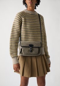 Maglione a righe giallo, grigio e beige con polsini a costine, abbinato a una gonna a pieghe verde oliva. Borsa a tracolla nera con design trapuntato.