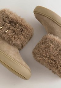 Bocanci din blană artificială bej, cu o textură moale, având o parte superioară din suede, o formă rotundă și accesorii din metal de culoare aurie. Talpa este din cauciuc plat.