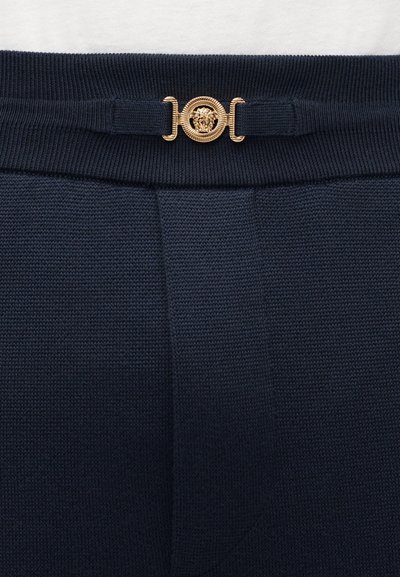 Marineblauwe gebreide broek met een gestructureerde stof, voorzien van een gouden leeuwenkopembleem op een decoratieve tailleband.