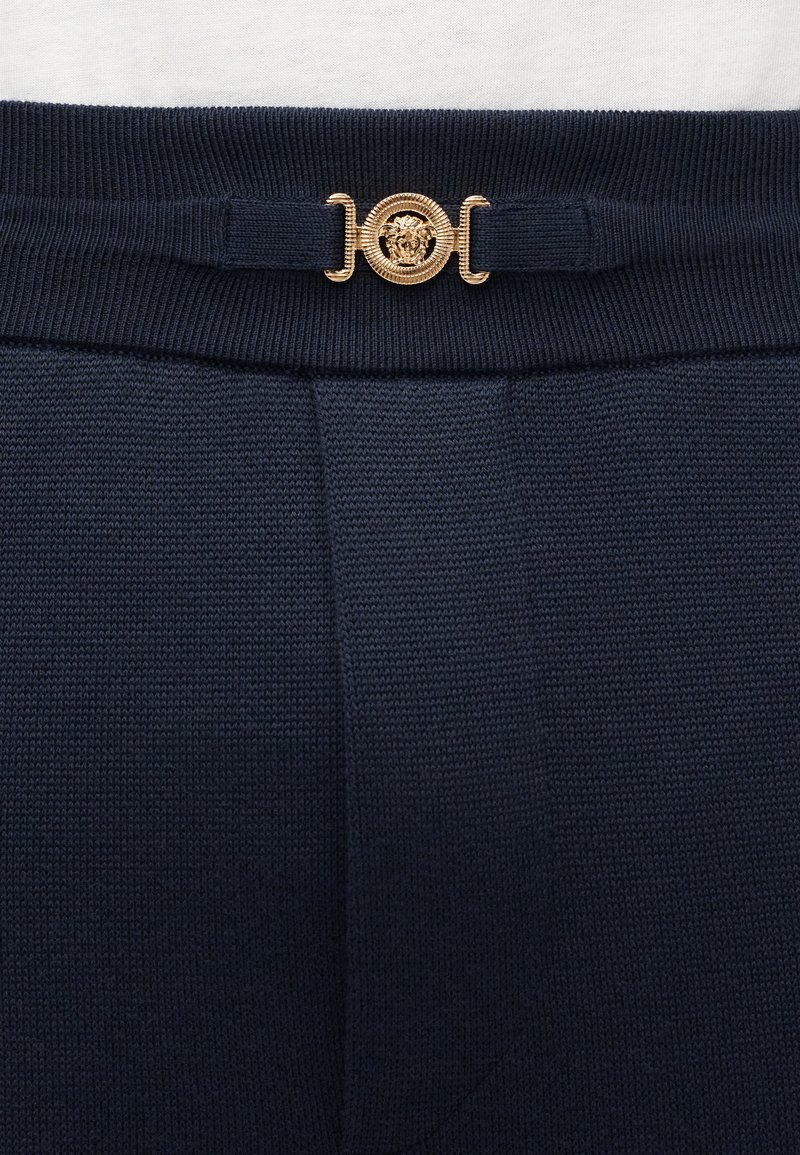 Marinblå stickade byxor med ett texturerat tyg, med en guldfärgad lejonhuvudemblem på en dekorativ midjeband.