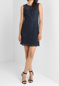 Granatowa koronkowa sukienka bez rękawów z dopasowaną górą i rozkloszowaną spódnicą. Połączona z czarnymi sandałami na szpilkach.