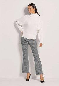 Weißer gestrickter Pullover mit hohem Kragen, kombiniert mit schwarz-weiß gemusterten Schlaghosen, akzentuiert durch schwarze High Heels.