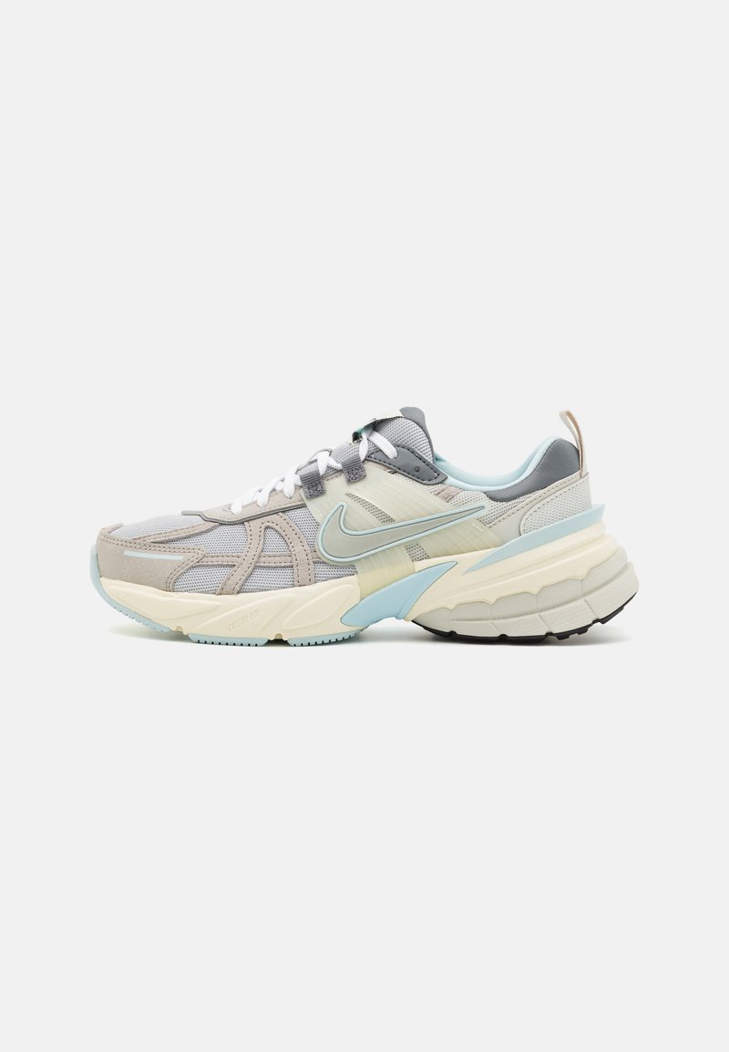 Zapatilla de running Nike en gris claro y beige con cordones blancos, suela acolchada y detalles en azul claro, vista de lado sobre un fondo blanco.