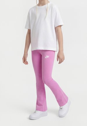 Personne portant des baskets blanches, des leggings roses avec le logo Nike, et un haut blanc ample à manches courtes, debout devant un fond blanc.