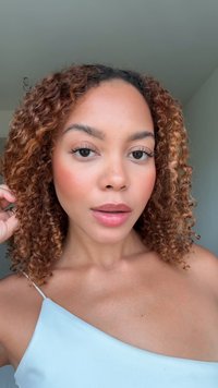 Lockar med en ljusbrun nyans, bär en ljusblå topp med spaghettiband. Har klar hud, neutral sminkning och en elegant pose.