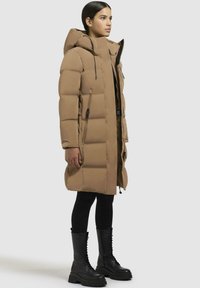 Brun pufferjakke med høj krave, lynlås foran og sidelommer; parret med sorte lårhøje snørebodystøvler og sorte leggings.